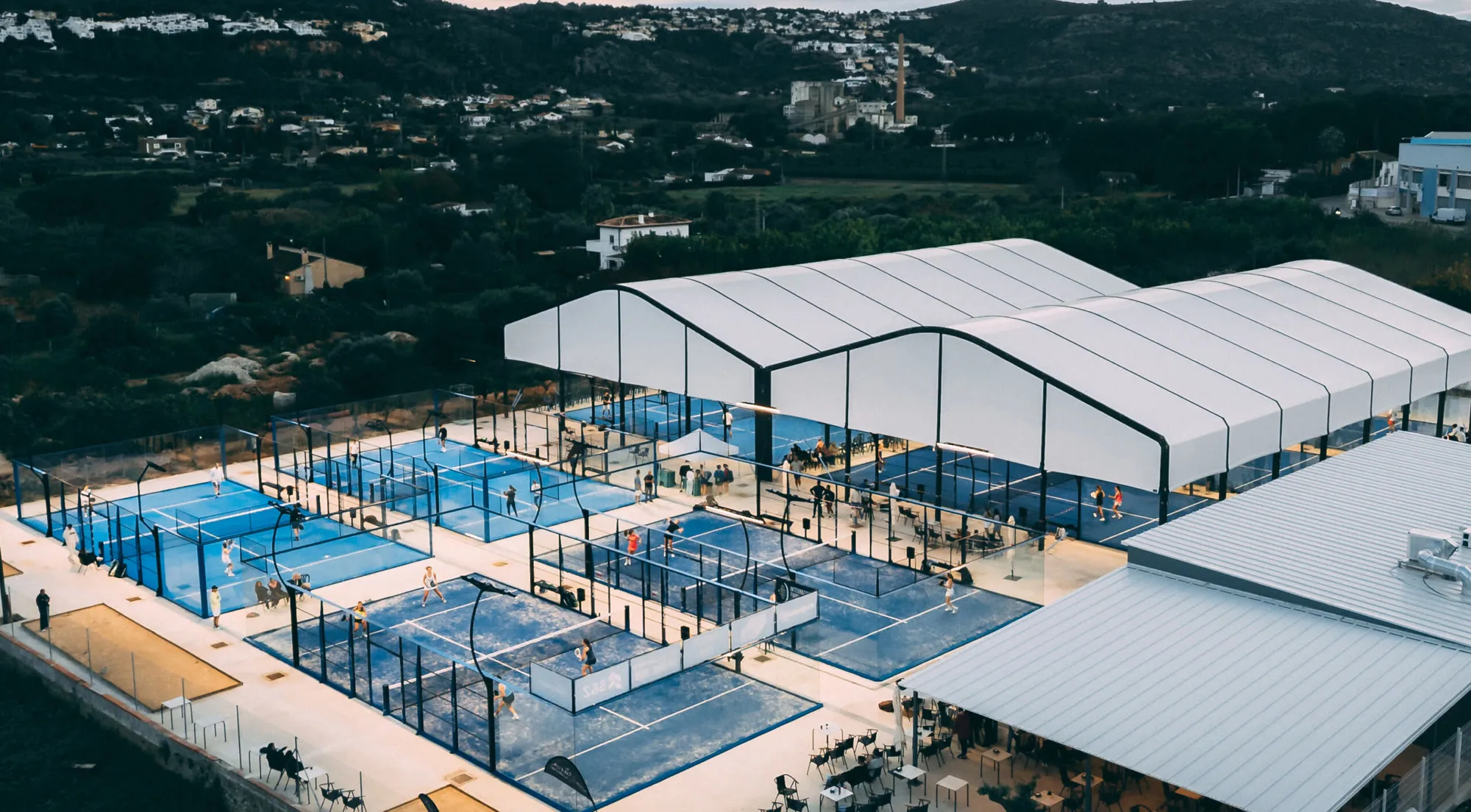 Luftbild der Anlage in der Dämmerung: acht Padelplätze, eine überdachte Halle und eine Caféterrasse, dahinter die Hügel der Marina Alta